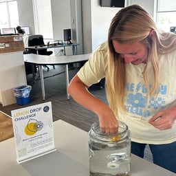 A person participates in the "Lemon Drop Challenge," attempting to balance a quarter on a lemon submerged in a clear jar of water.