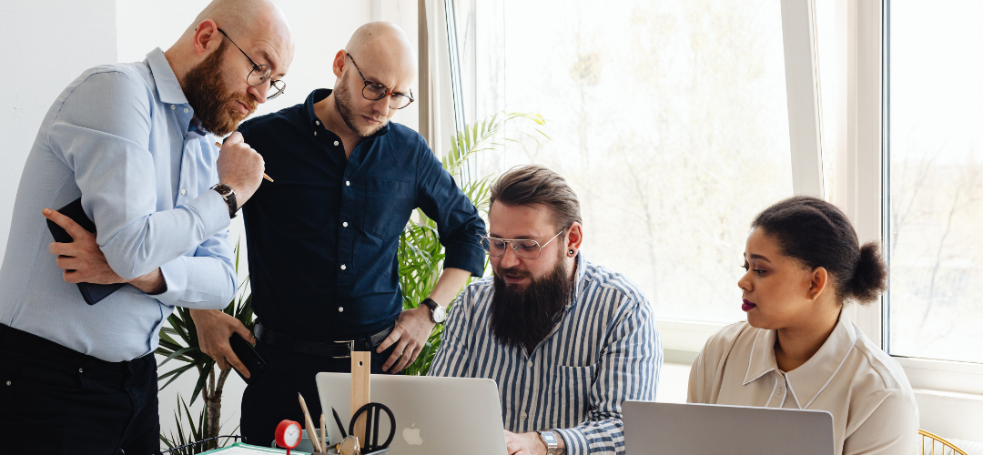A group of four professionals collaborate around a table, focused on laptops in a bright office with greenery in the background.