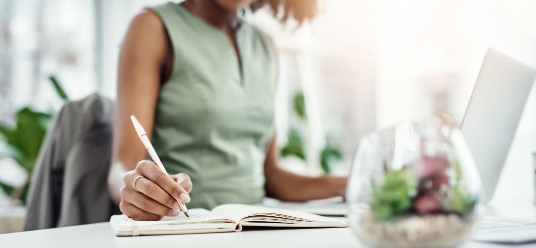 A person in a green top writes in a notebook beside a laptop and a small plant terrarium, focused on their work.