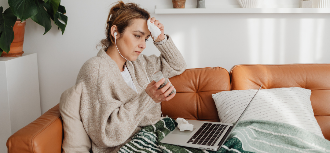 A person sits on a couch with a blanket, holding a jar and resting a hand on their forehead, with a laptop in front of them.