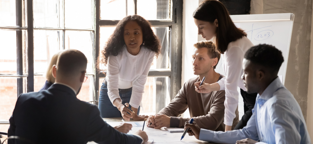 A diverse team collaborates around a table, engaged in discussion and note-taking, with a whiteboard in the background.