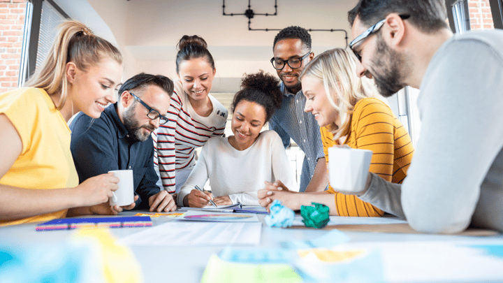 A diverse group of people collaborates at a table filled with papers and coffee mugs, brainstorming ideas in a modern office space.