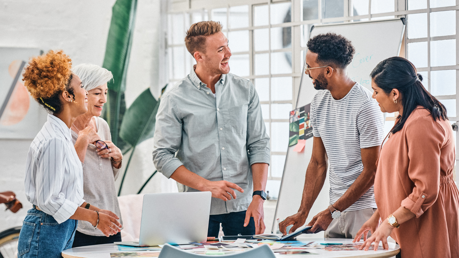 A diverse group of professionals collaborates around a table filled with colorful design materials and a laptop in a bright office space.
