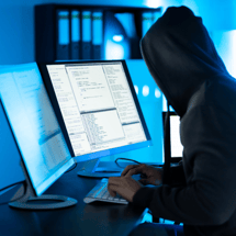 A hooded figure types on a keyboard in a dimly lit room filled with glowing monitors displaying lines of code.