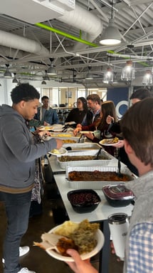 A group of people gathered around a buffet table filled with various dishes, serving food onto their plates in a modern dining space.