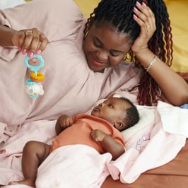 A mother holds a colorful baby toy while lying next to a peaceful baby swaddled in soft blankets in a cozy setting.
