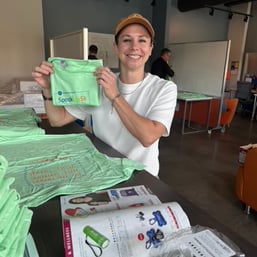 A person holds a green t-shirt branded "SpeakUp5k" amidst a stack of similar shirts on a table in a casual workspace.