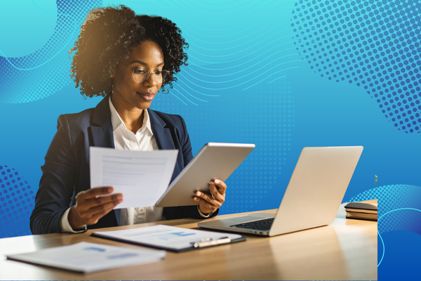A person in a suit holds documents and a tablet while seated at a desk with a laptop, against a vibrant blue background.