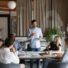 A group meeting in a modern office, with one person presenting ideas to attentive colleagues around a table filled with documents and stationery.