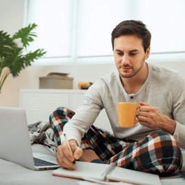 A person sits on a bed in casual attire, holding a mug and writing notes beside a laptop, surrounded by a bright, airy room.