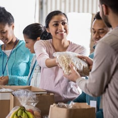 A woman is receiving a bag of food from another person at a food distribution event, surrounded by others waiting in line.