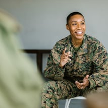 A U.S. Marine in camouflage uniform gestures while engaged in discussion, seated in a spacious, minimalistic room.
