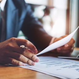 A professional in a suit examines documents, holding a pen in one hand and papers in the other, focused on the task at hand.
