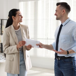 A woman in a beige blazer accepts an envelope from a man in a light blue shirt and black tie in a bright room.