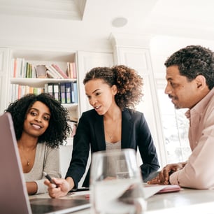 Three individuals engage in a discussion around a table, with a laptop and a glass of water nearby. Books are visible on a shelf.