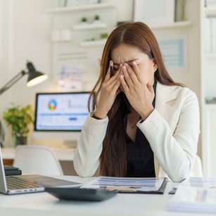 A woman in a white blazer sits at a desk with papers and a laptop, appearing stressed while holding her head with both hands.