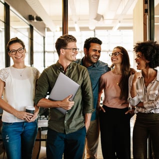 A group of five professionals stands together in a modern office setting, engaged in conversation and exuding a collaborative atmosphere.