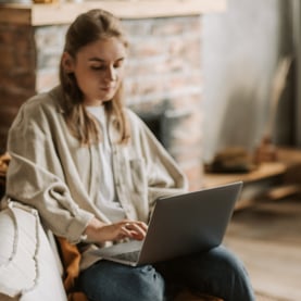 A person sits on a couch, focused on a laptop, wearing a cozy sweater, in a stylish, minimalistic room with a brick wall.
