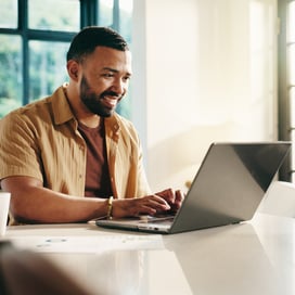 A person in a beige shirt sits at a table, typing on a laptop with sunlight streaming in through large windows.