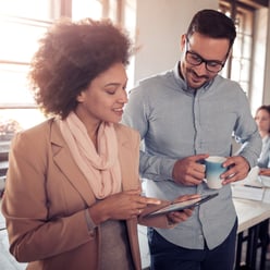 A woman in a beige blazer and scarf looks at a tablet while a man in a light blue shirt holds a cup, both engaged in conversation.